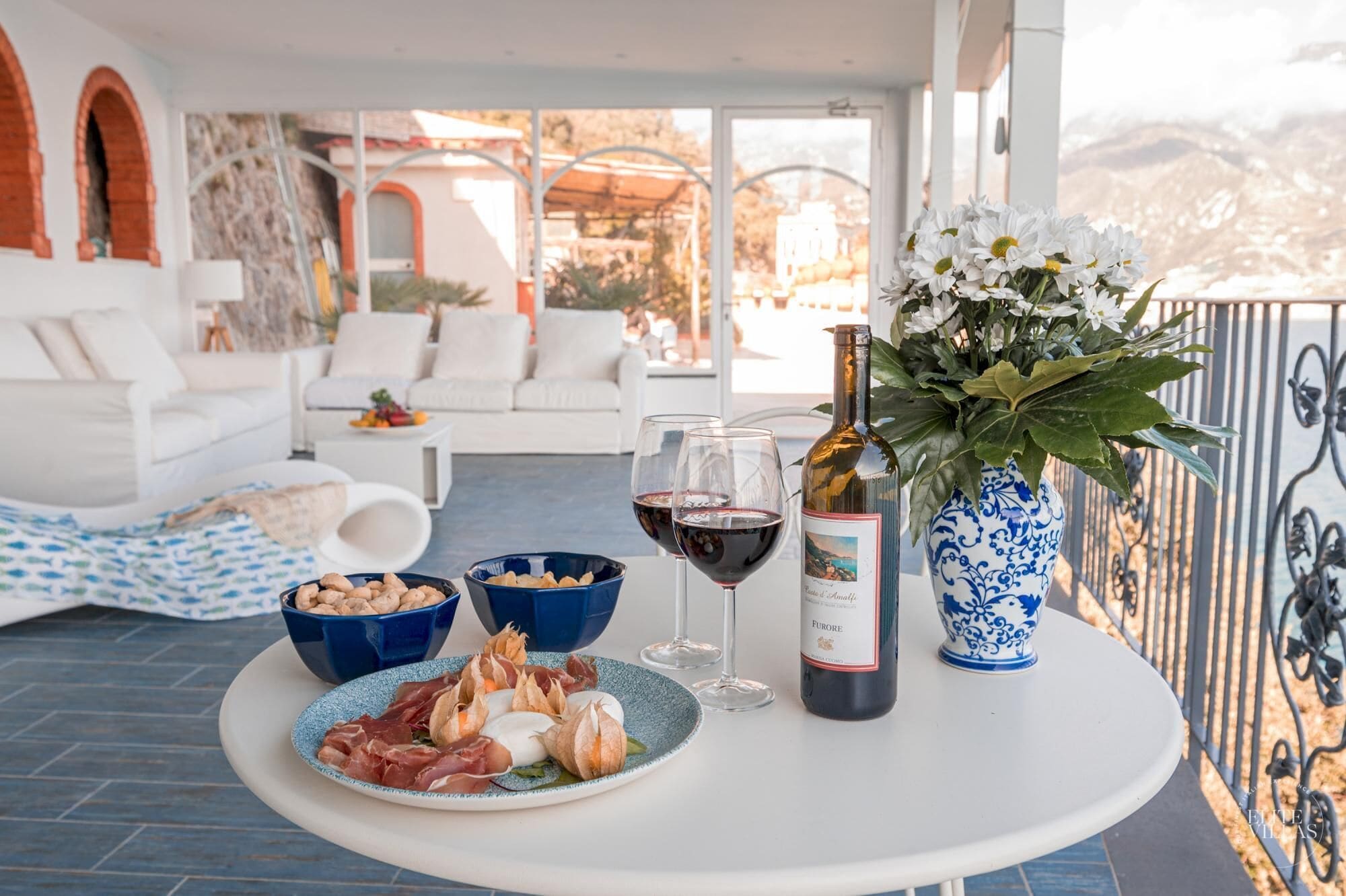 Une terrasse avec une table dressée pour un repas, comprenant une bouteille de vin, un vase de fleurs et une assiette de nourriture.