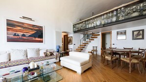 En rummelig opholdsstue med sofa, sofabord i glas og spisebord med stole. Der er en trappe med glasgelænder, som fører til en etage ovenover.