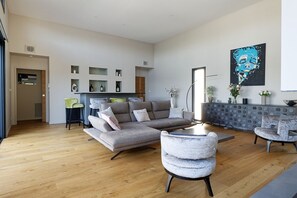 Een ruime woonkamer met een grijze hoekbank, een ronde salontafel en een grijze fauteuil. Er is een moderne haard met decoratieve schouw, en een groot abstract schilderij aan de muur.