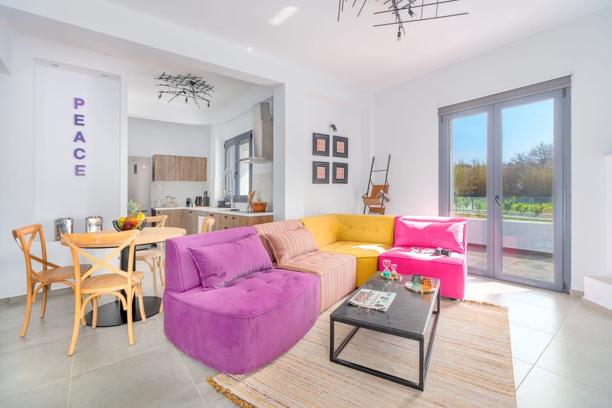 Un salon moderne avec un canapé violet, un canapé d’angle jaune et un fauteuil rose. On aperçoit une table à manger avec des chaises et une cuisine au fond.