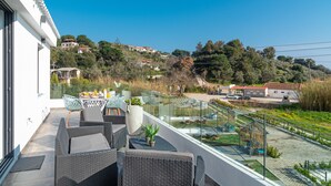 Un balcone con mobili in vimini, ringhiera in vetro e vista su una collina con edifici.