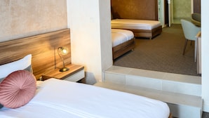 A hotel room with a bed, a nightstand with a lamp, a chair, and a small desk. - Monogram (Inđija)