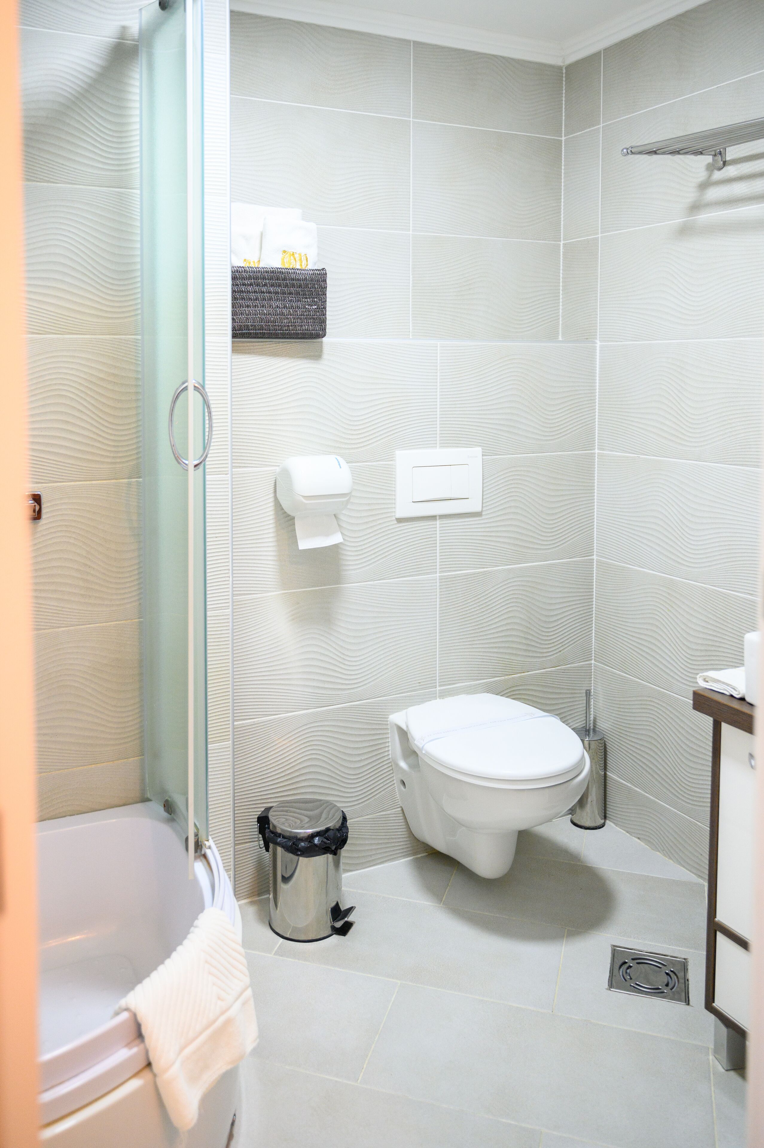 A bathroom with a toilet, a bidet, a glass shower enclosure, a trash bin, and a towel rack.