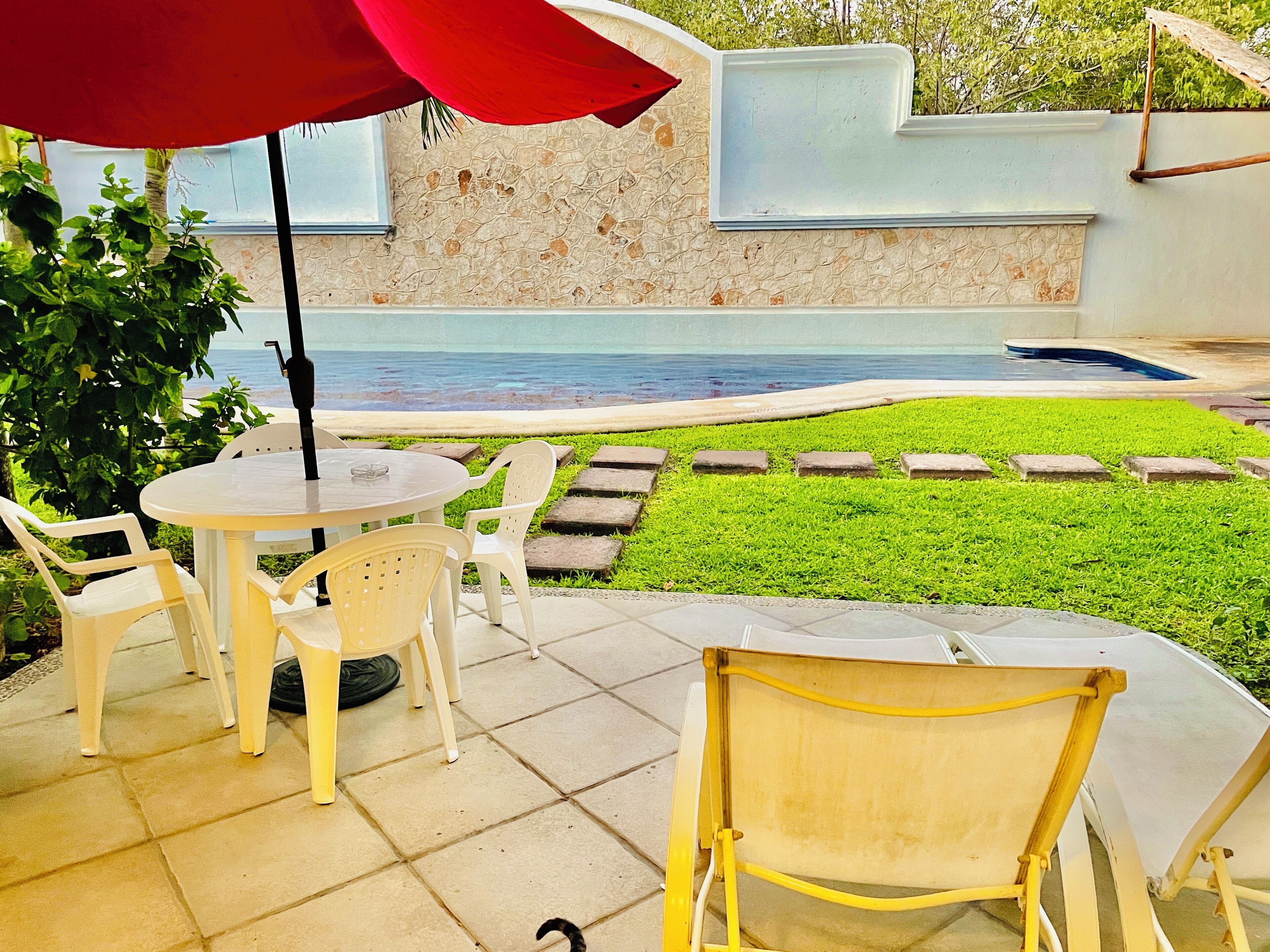 A patio area with a round table and chairs, a red umbrella, and a view of a swimming pool.