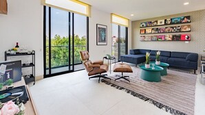 Un salon moderne avec un grand canapé, une table basse et un fauteuil. On y trouve une bibliothèque remplie de divers objets et une fenêtre donnant sur un paysage verdoyant.