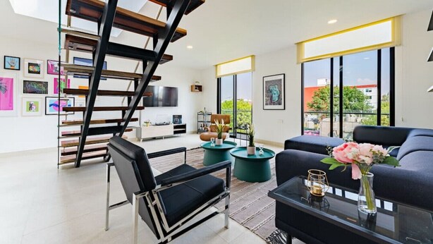 Un salon moderne avec un canapé noir, une table basse en verre et un fauteuil noir. On y trouve un escalier avec des poutres en bois et un téléviseur fixé au mur.
