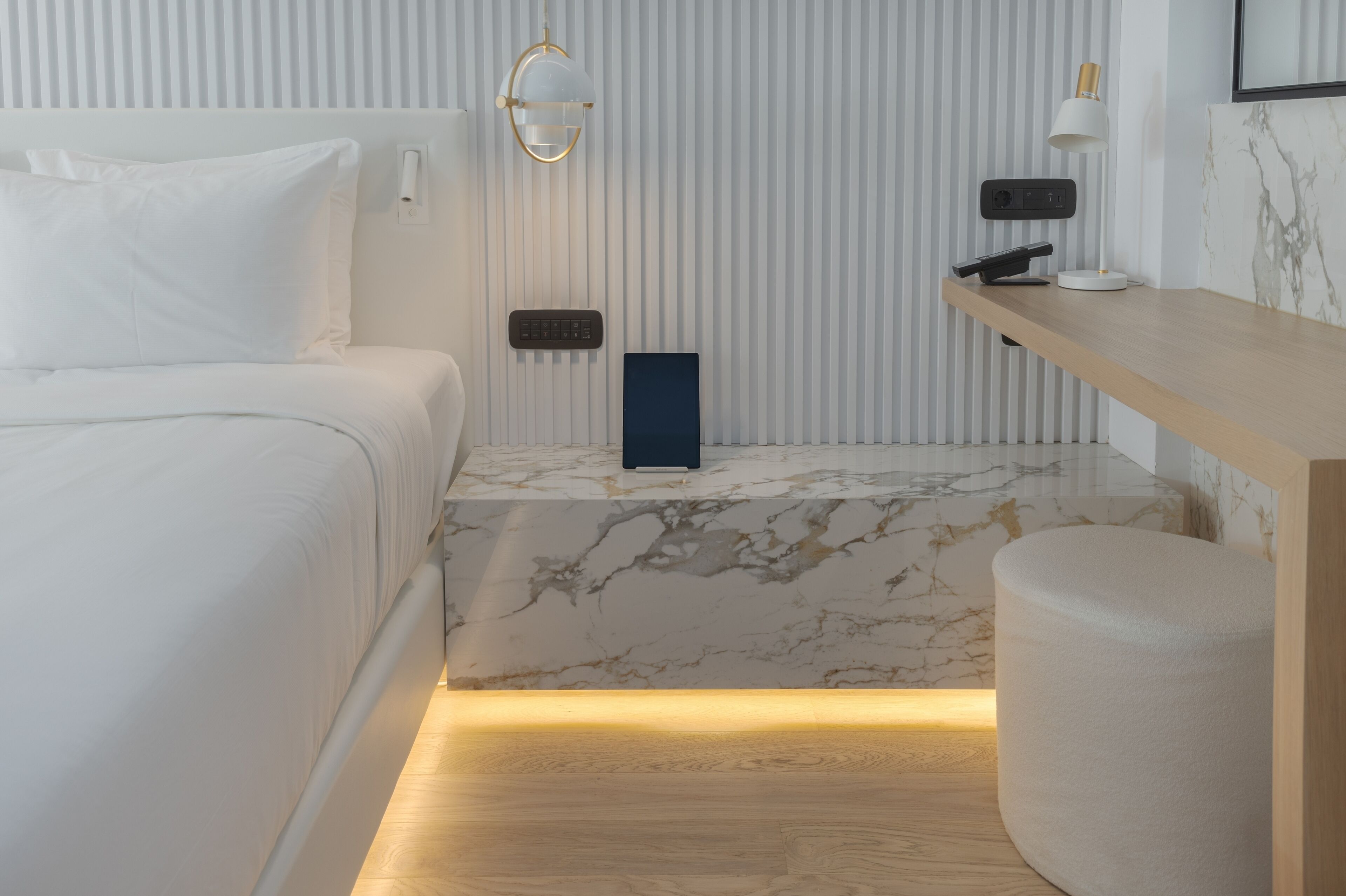 A modern hotel room with a bed, a bedside table with a lamp, a small blue object on the nightstand, and a marble-patterned wall.