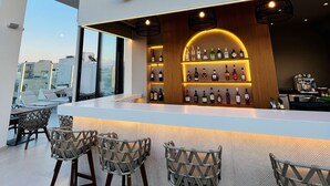 Un bar situé sur un toit, avec un panneau lumineux au néon, des tabourets de bar et une variété de bouteilles d’alcool.