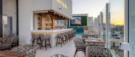 A rooftop bar with a neon sign, outdoor seating, and a large screen displaying a scenic view.