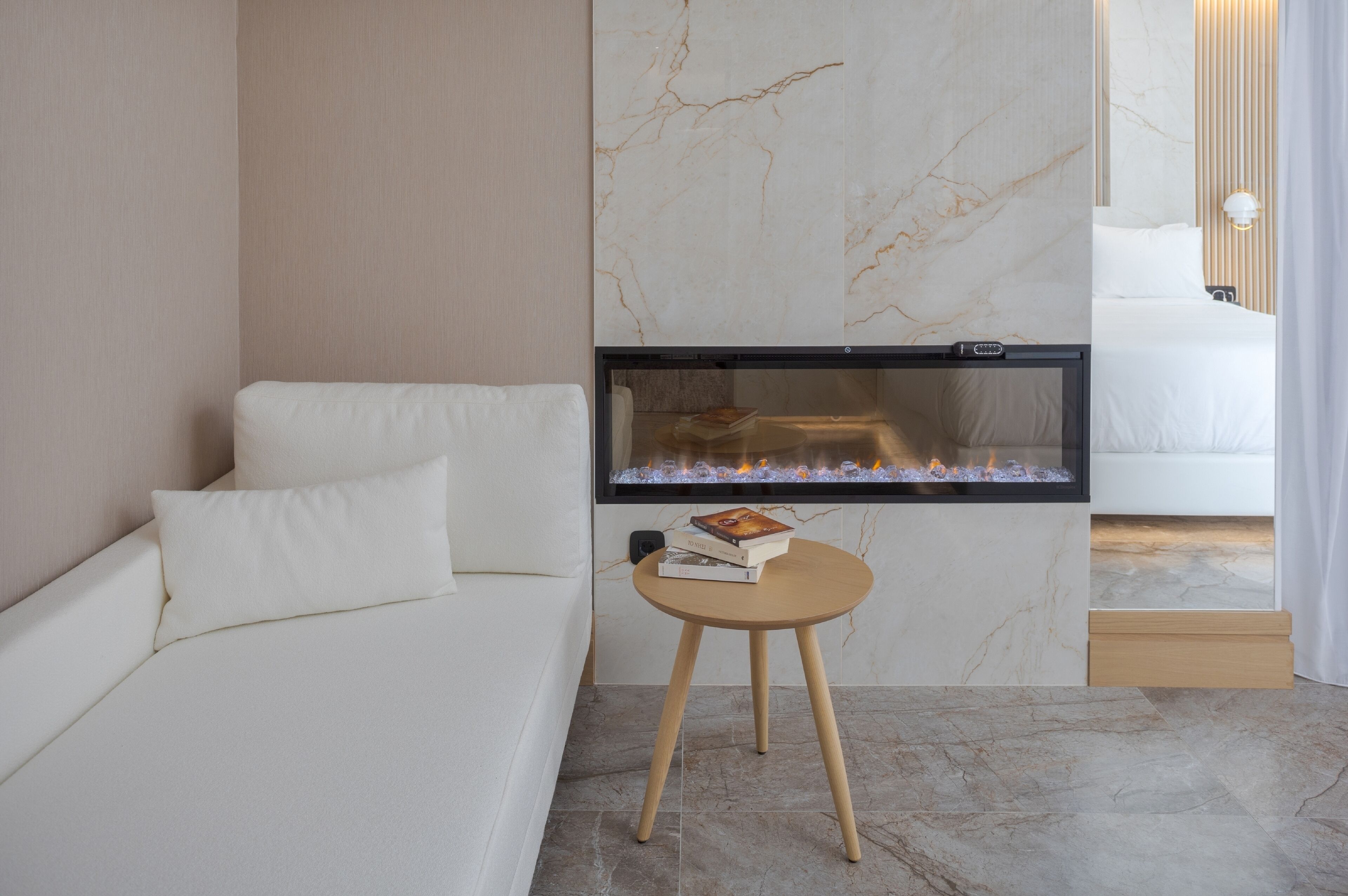 A modern hotel room with a white sofa, a small wooden table with a book, and a large wall-mounted fireplace.