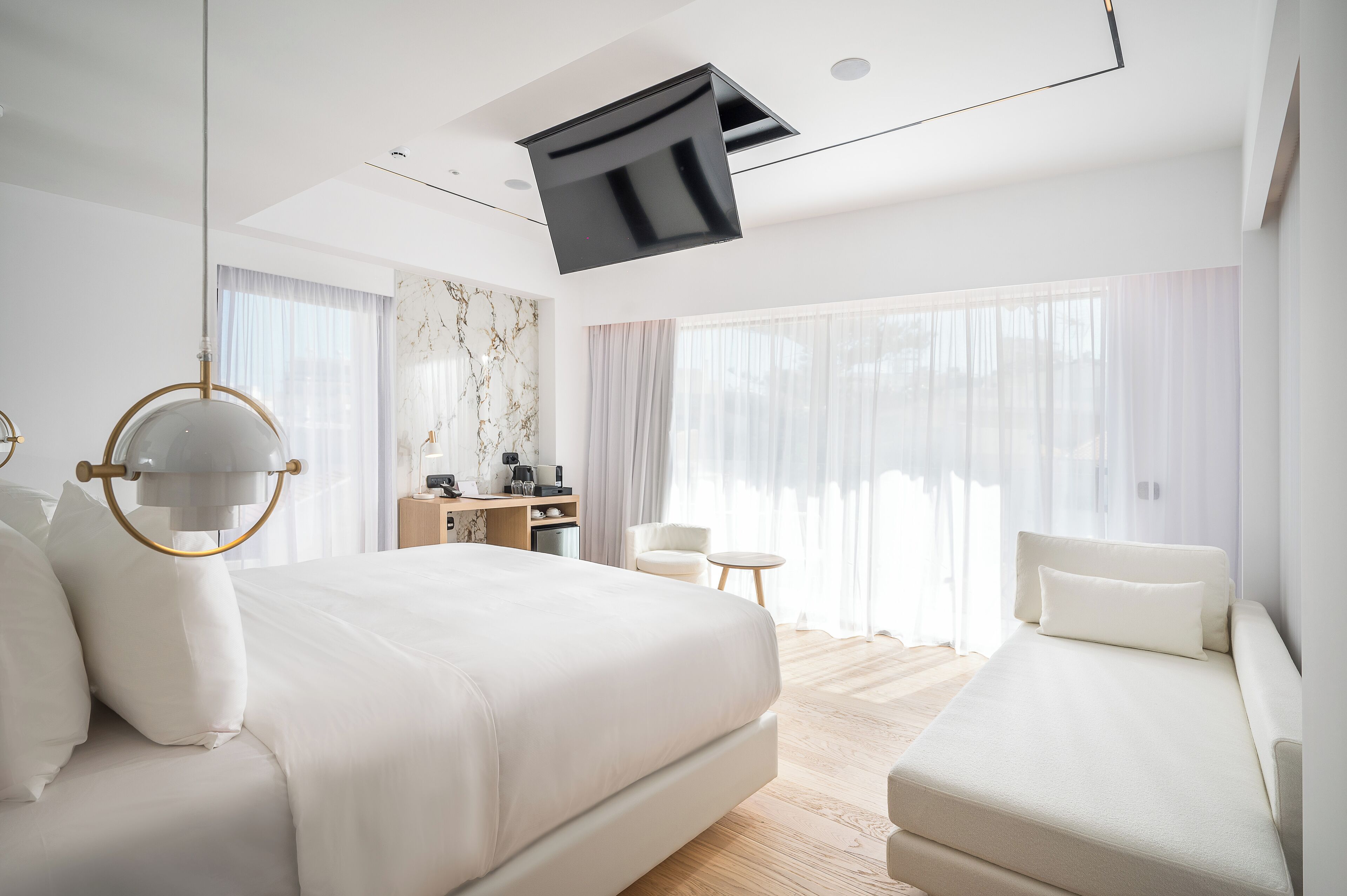 Une chambre d’hôtel moderne, dotée d’un grand lit, d’un canapé, d’un bureau et d’un téléviseur fixé au plafond.