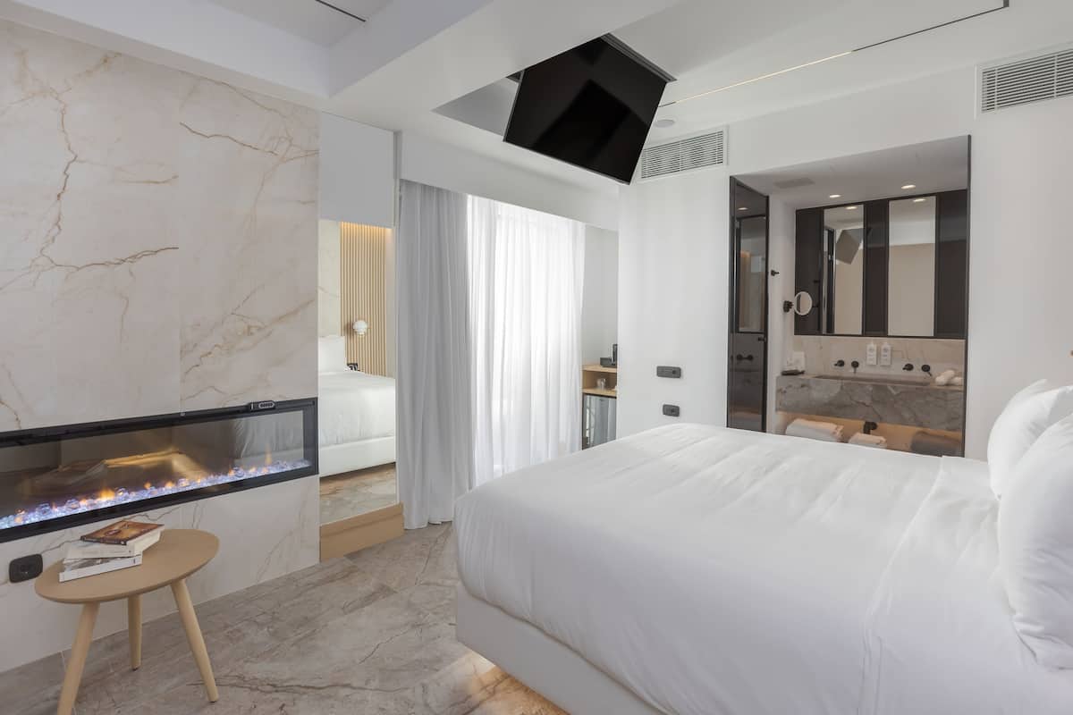 Une chambre d’hôtel moderne dotée d’un grand lit, d’une cheminée, d’une petite table avec des livres et d’une salle de bain comprenant un miroir et un lavabo.