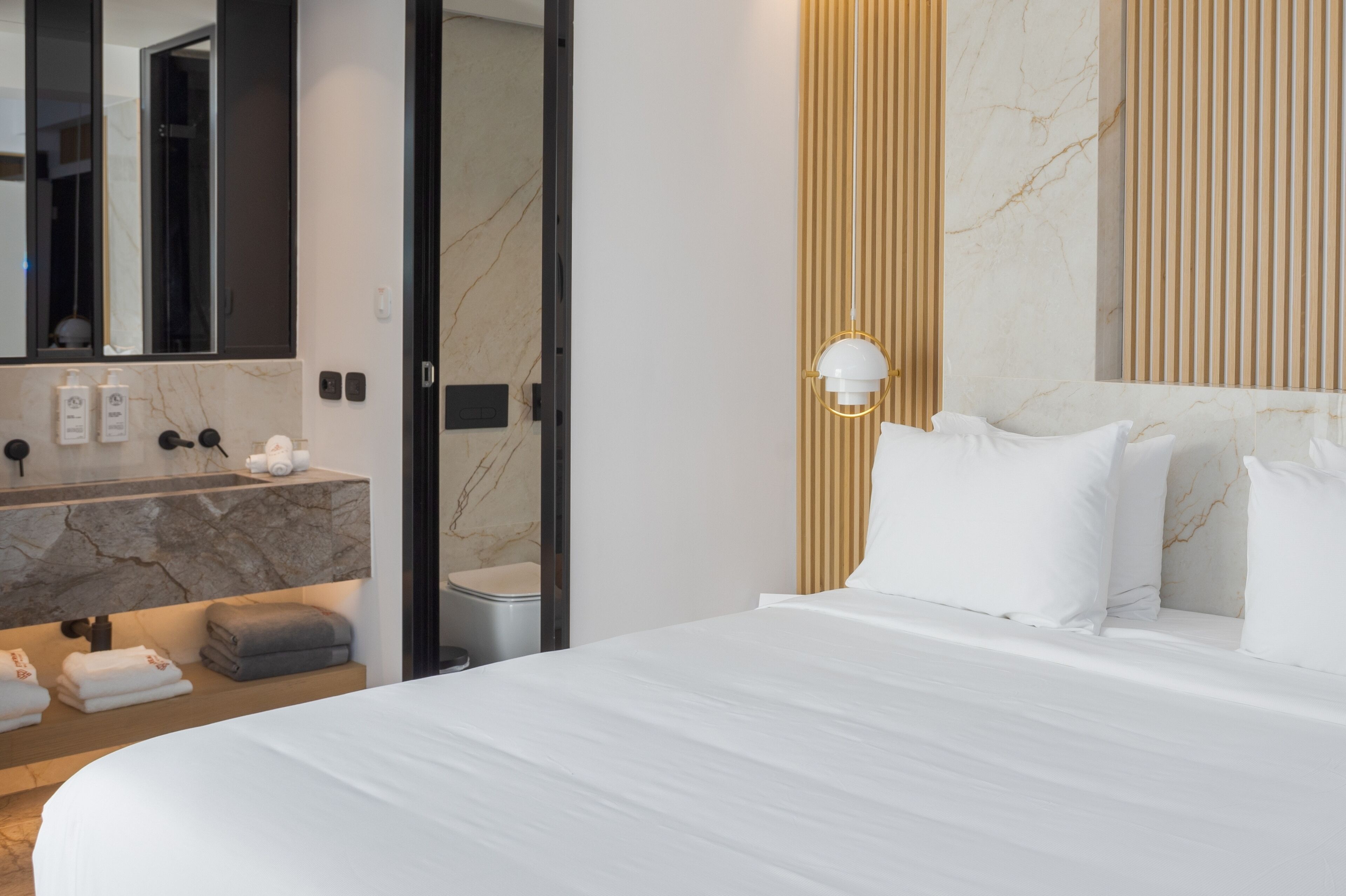 A hotel room with a large bed, a marble bathroom vanity, a mirror, and a wall-mounted light fixture.