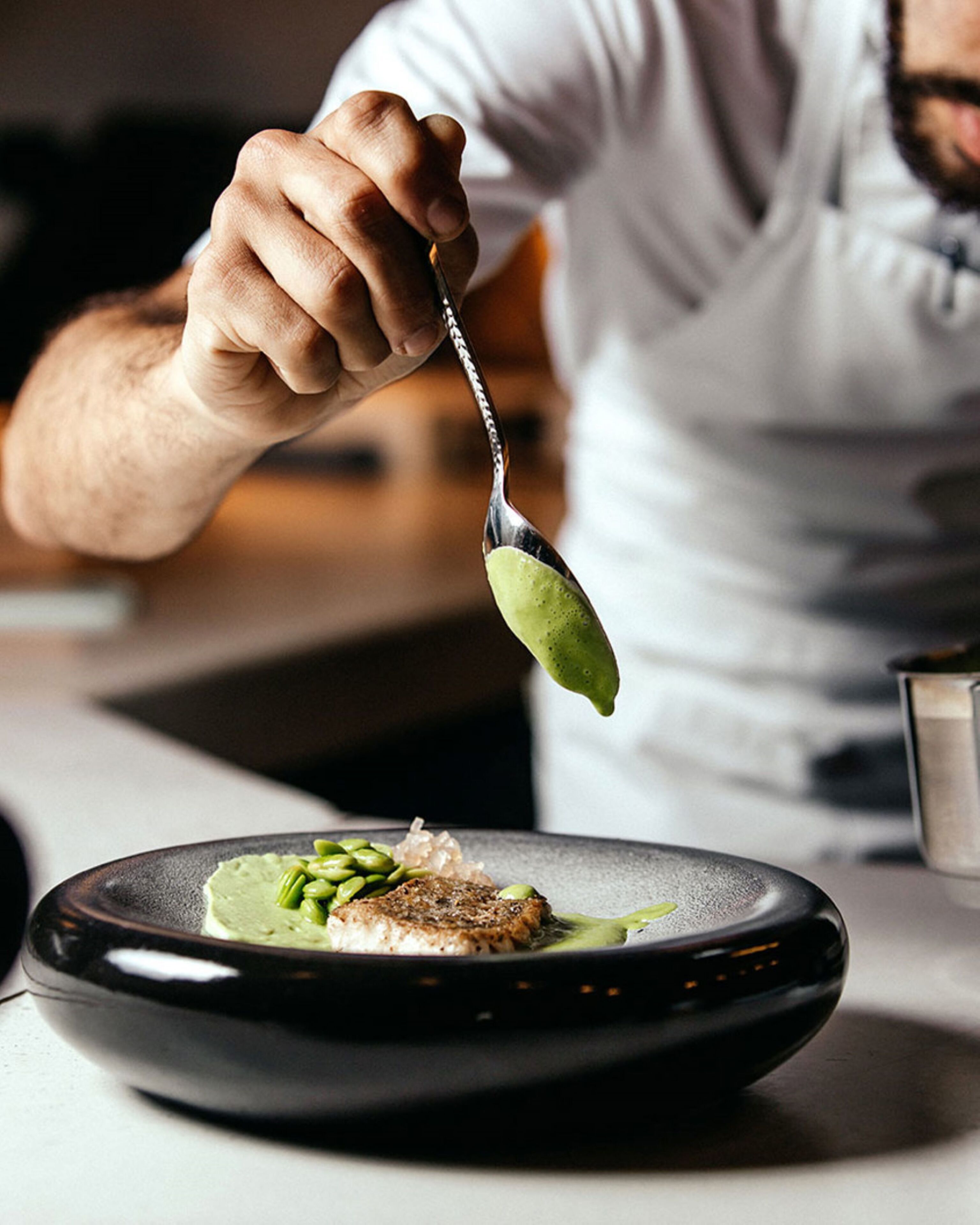 Une personne tenant une cuillère pleine d’une sauce verte, une assiette avec de la nourriture et un verre d’eau.