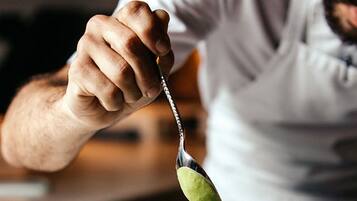 A person holding a spoon with a green sauce, a plate with food, and a glass of water.