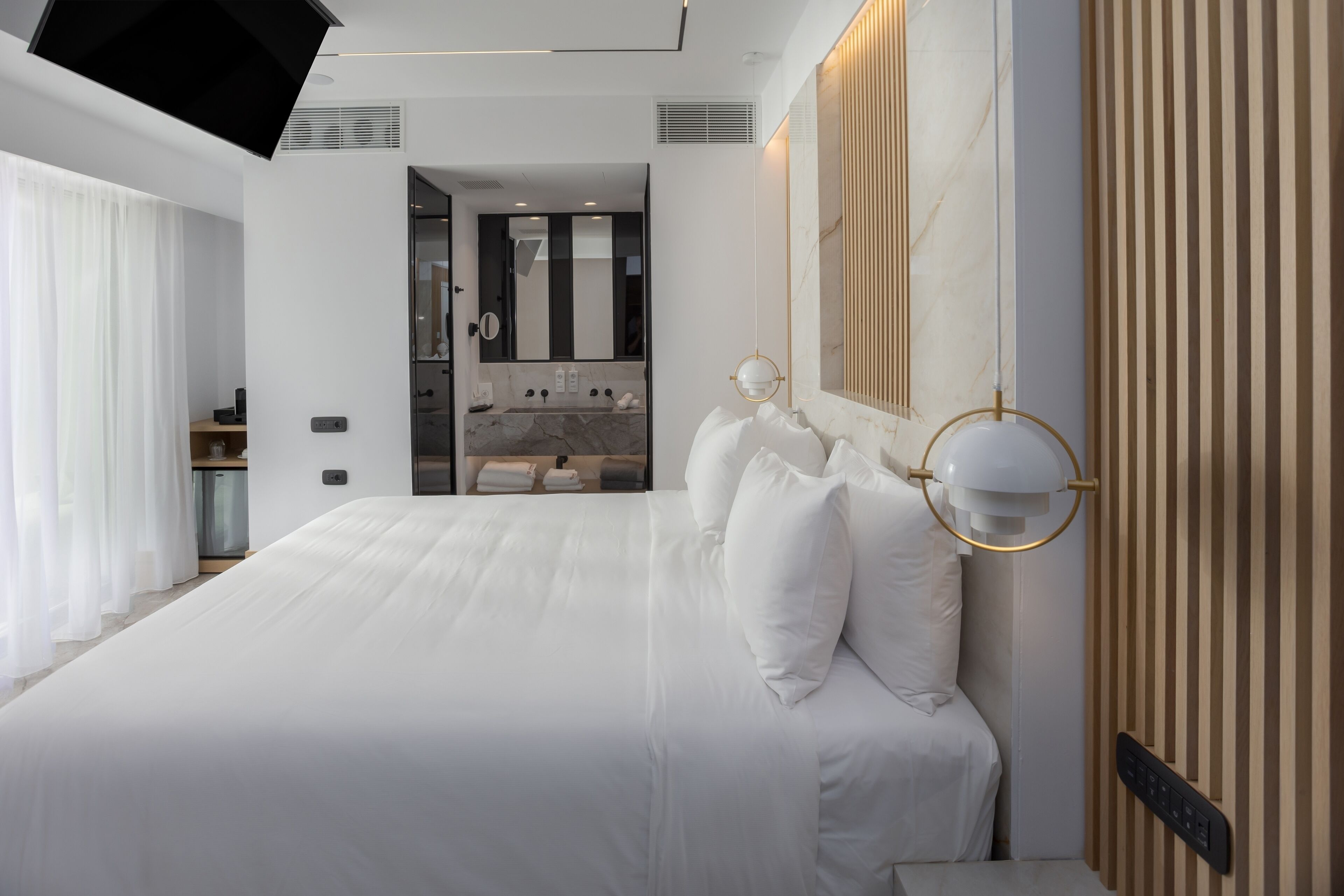 A modern hotel room with a large bed, a bathroom with a mirror and sink, and a TV mounted on the ceiling.