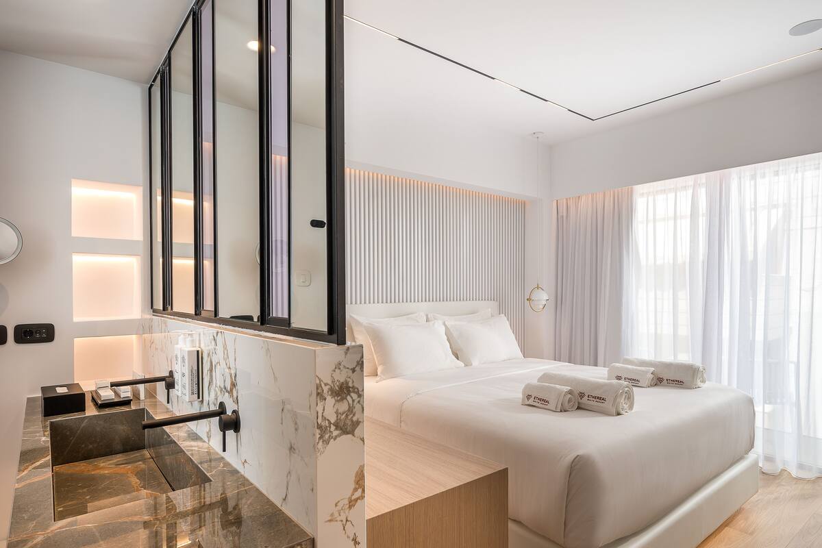 Une chambre d’hôtel moderne dotée d’un grand lit, d’un meuble-lavabo en marbre et d’une grande fenêtre agrémentée de rideaux légers.