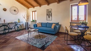 Ein Wohnzimmer mit einem blauen Sofa, einem Esstisch mit Stühlen, einem Fahrrad und einem großen Fenster mit Blick auf ein Dach.