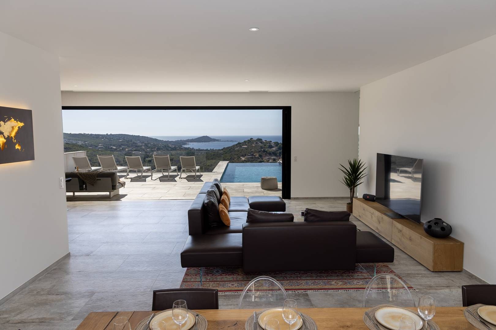 A modern living room with a large window, a sofa, a TV, and a dining area with a view of the sea.