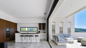Una cocina moderna con barra, zona de comedor con sofá y reposapiés, y balcón con vistas a las colinas.