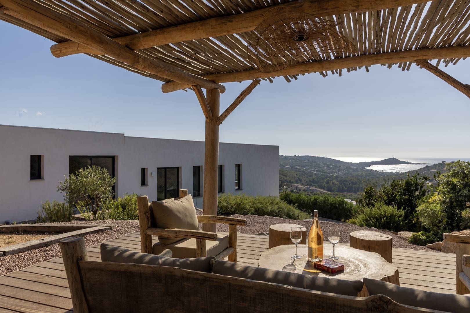 Un patio al aire libre con mobiliario de madera, una mesa con vasos y vistas a un edificio moderno y un paisaje.