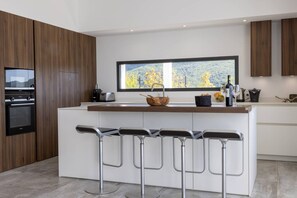 Een moderne keuken met een centraal eiland, barstoelen, een inbouwoven en een groot raam met uitzicht.