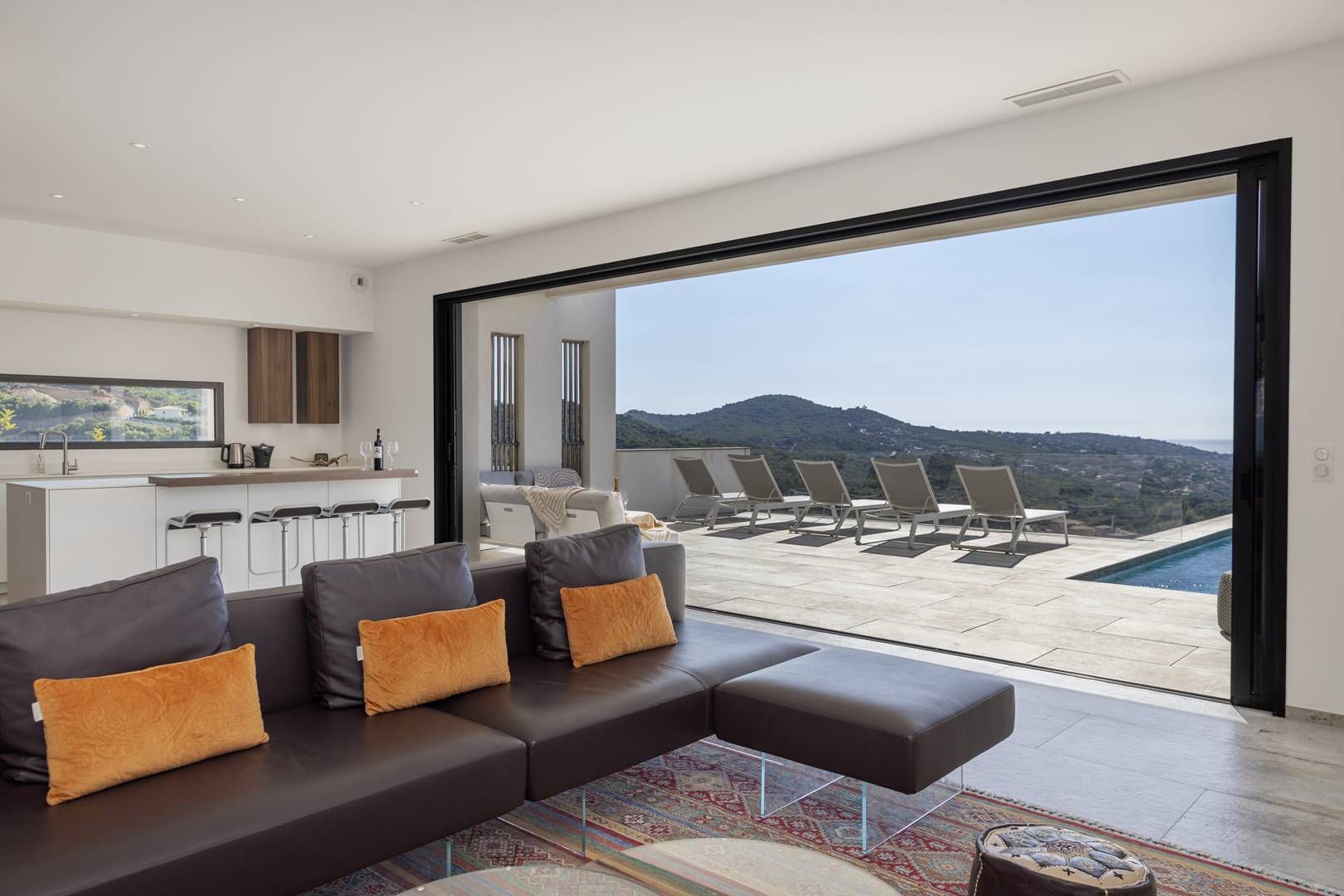 Een moderne woonkamer met een bank, een keukenhoek en een balkon met een zwembad en stoelen.