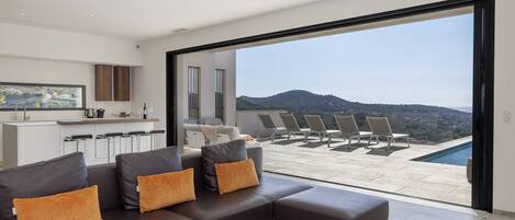 A modern living room with a sofa, a kitchen area, and a balcony with a pool and chairs.