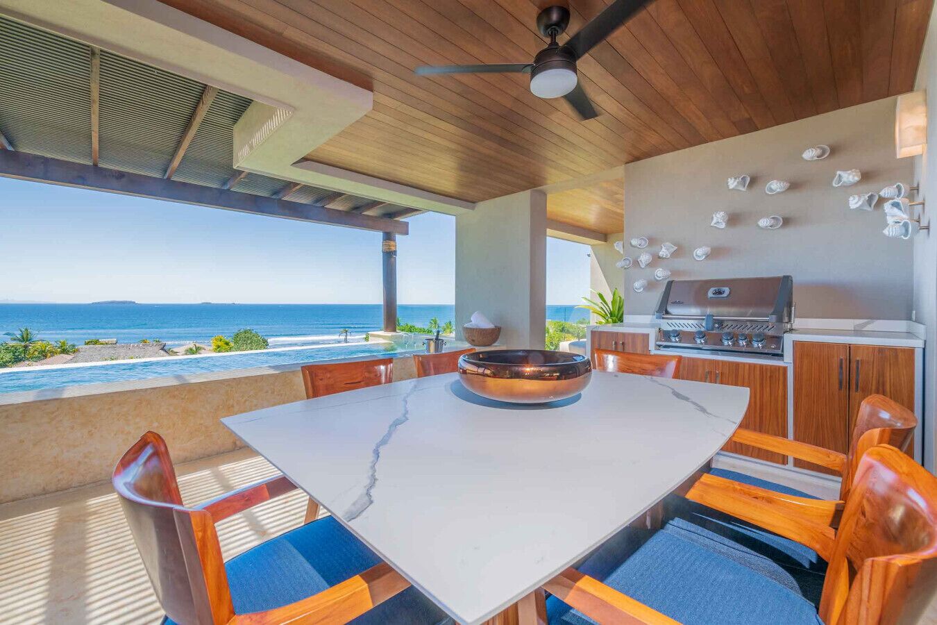 Une cuisine extérieure moderne avec vue sur l’océan, une table à manger et un ventilateur de plafond.