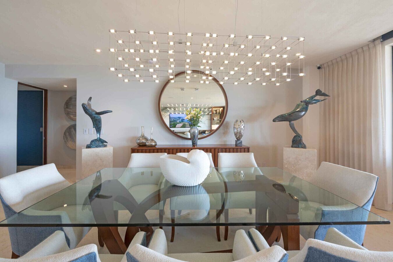 Une salle à manger moderne avec une table en verre, des chaises blanches, un grand miroir central et des sculptures décoratives.