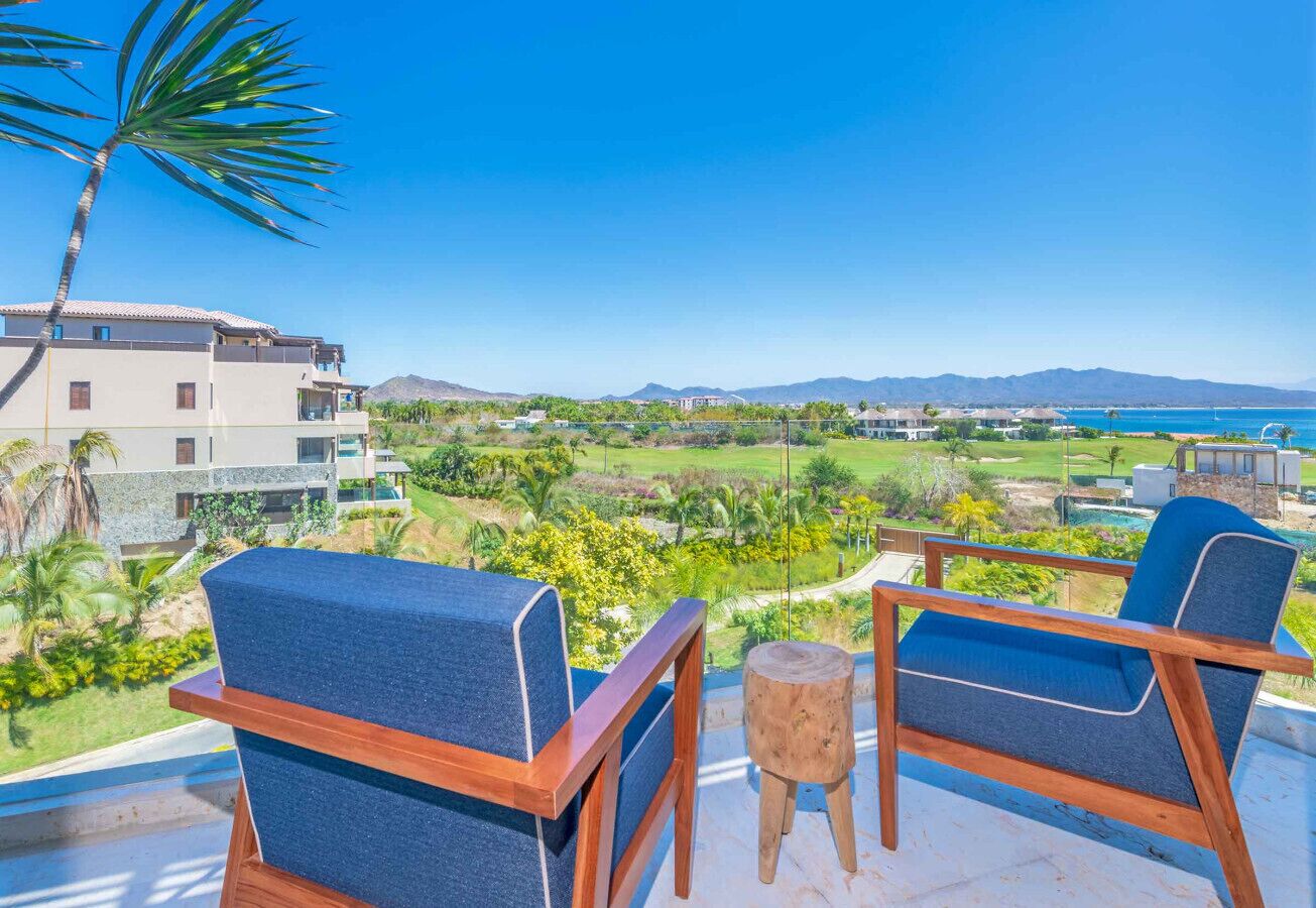 A balcony with two blue chairs and a small wooden stool, overlooking a landscaped garden and a distant view of mountains and a beach.