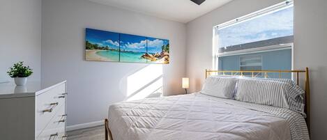 Une chambre à coucher avec un lit, un ventilateur de plafond, une fenêtre, une table de chevet et un tableau représentant une scène de plage.