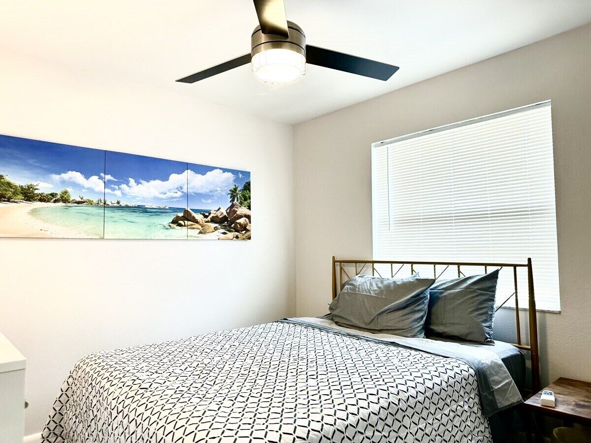 A bedroom with a bed, a ceiling fan, a window with blinds, and a beach-themed wall art.