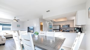 Une cuisine moderne avec des meubles blancs, un coin repas avec une table en bois et des chaises blanches, et un salon avec un canapé et un ventilateur de plafond.