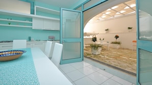 A modern kitchen with white cabinets, a dining table with a blue patterned table runner, and a view of an outdoor area with potted plants. - 3241 Villa Esador by Barbarhouse (Racale)