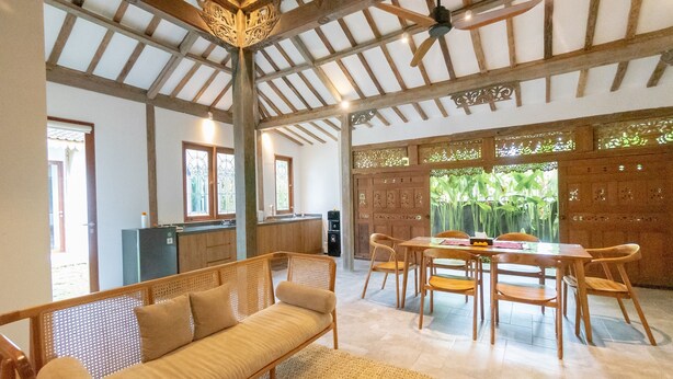 A spacious living area with a high ceiling, wooden beams, a dining table with chairs, a sofa, and a kitchen in the background. - Singgah di Ubud 1 (Ubud)