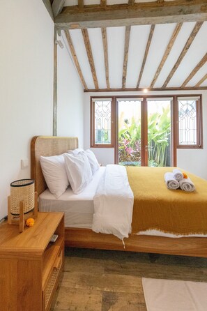 A bedroom with a wooden bed, white bedding, a wooden nightstand, and a view of greenery through a window. - Singgah di Ubud 1 (Ubud)