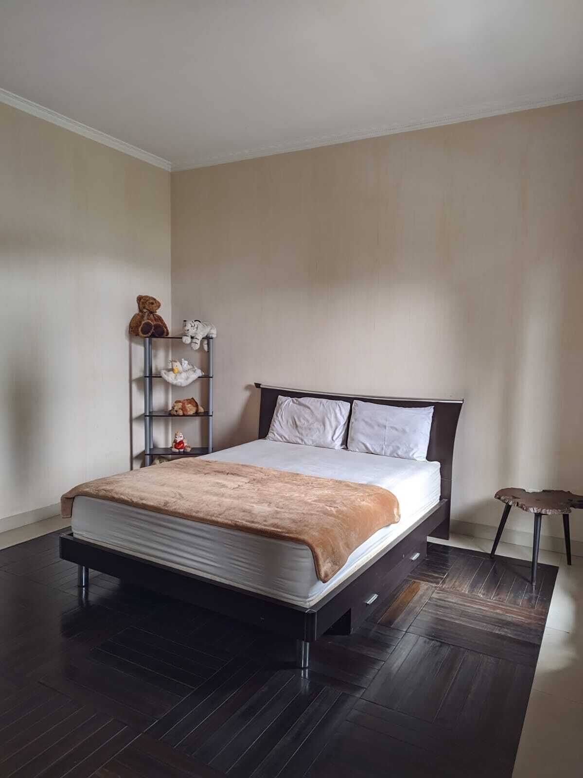 Un dormitorio moderno con piso de madera oscura, una cama con colchón blanco y manta marrón, un estante metálico con juguetes de peluche y una pequeña mesa redonda.
