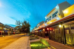 A well-lit street with buildings, a paved walkway, and a small garden area. - Hoi An Town Home Resort (Da Nang)