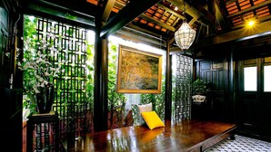 A traditional room with a wooden floor, a bed with a yellow pillow, a painting on the wall, and a window with a view of greenery. - Hoi An Town Home Resort (Da Nang)
