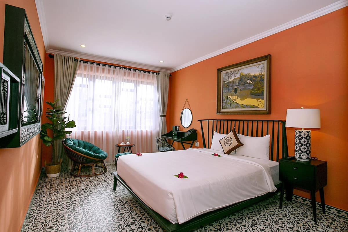 A hotel room with a bed, a chair, a desk, a lamp, and a painting on the wall.