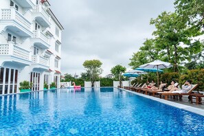 A swimming pool with lounge chairs and umbrellas, surrounded by a white building with balconies. - Hoi An Town Home Resort (Da Nang)