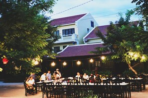 An outdoor dining area with tables and chairs, surrounded by trees and a building in the background. - Hoi An Town Home Resort (Da Nang)