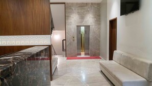 A modern hotel lobby with a marble reception desk, a sofa, and a marble floor leading to an elevator. - Pine Regency - Aravali's Peak view (Udaipur)