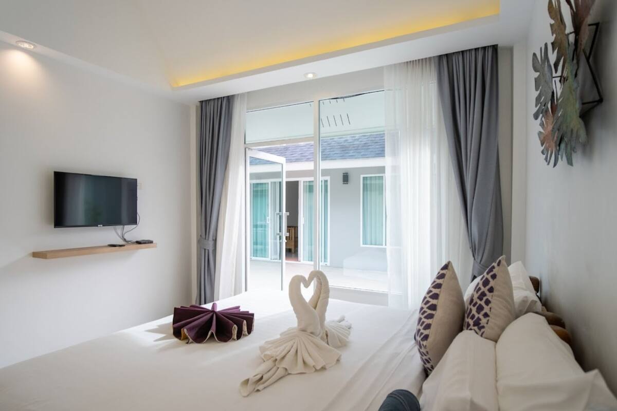 A modern hotel room with a flat-screen TV, a bed with white bedding, two swan-shaped towels, and a view of a building through large windows.