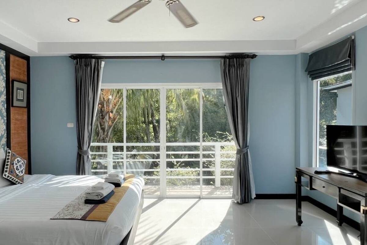A bedroom with a bed, a TV, a window with curtains, and a ceiling fan.
