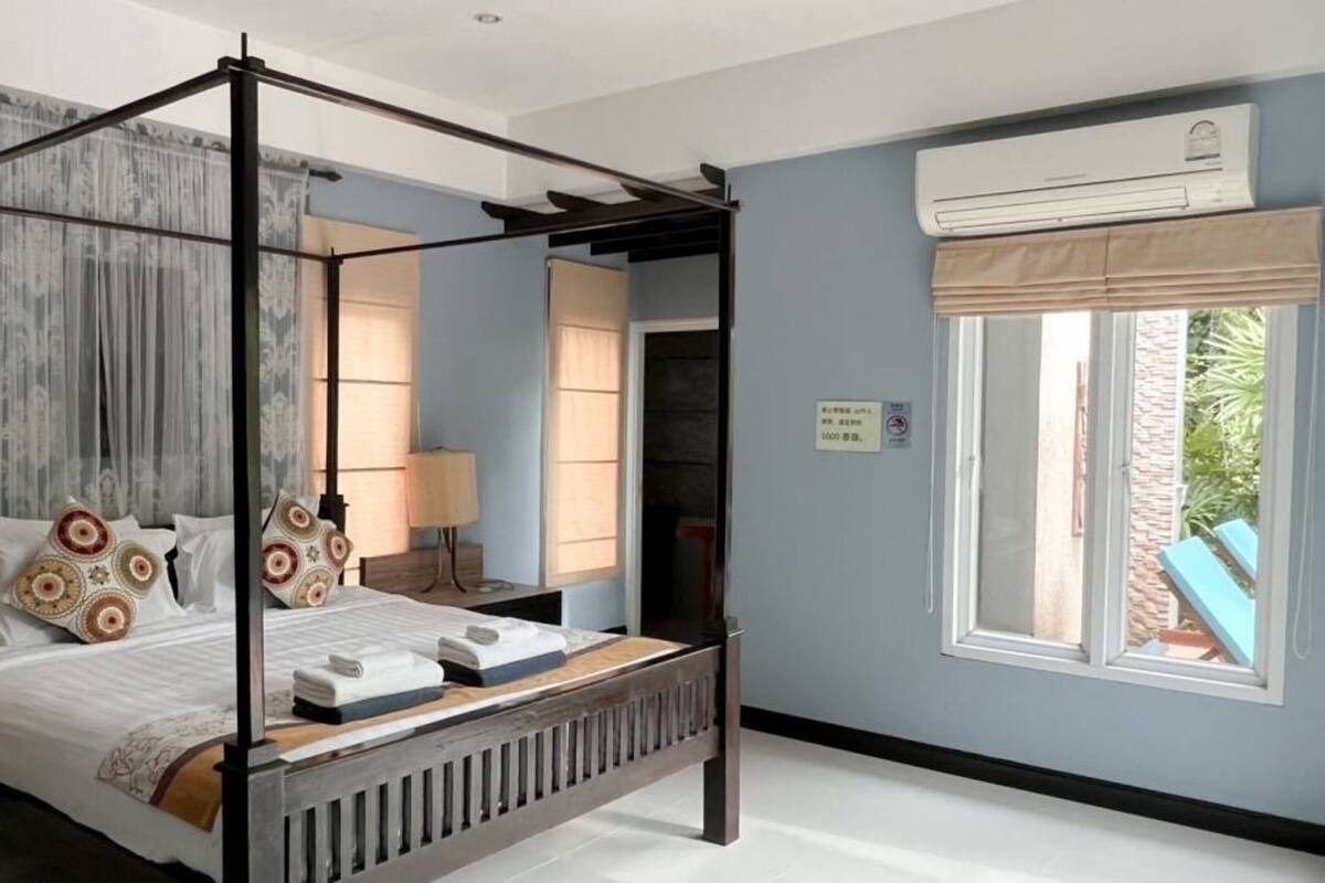 A four-poster bed with a canopy, a bedside table, a lamp, and a window with an air conditioning unit.