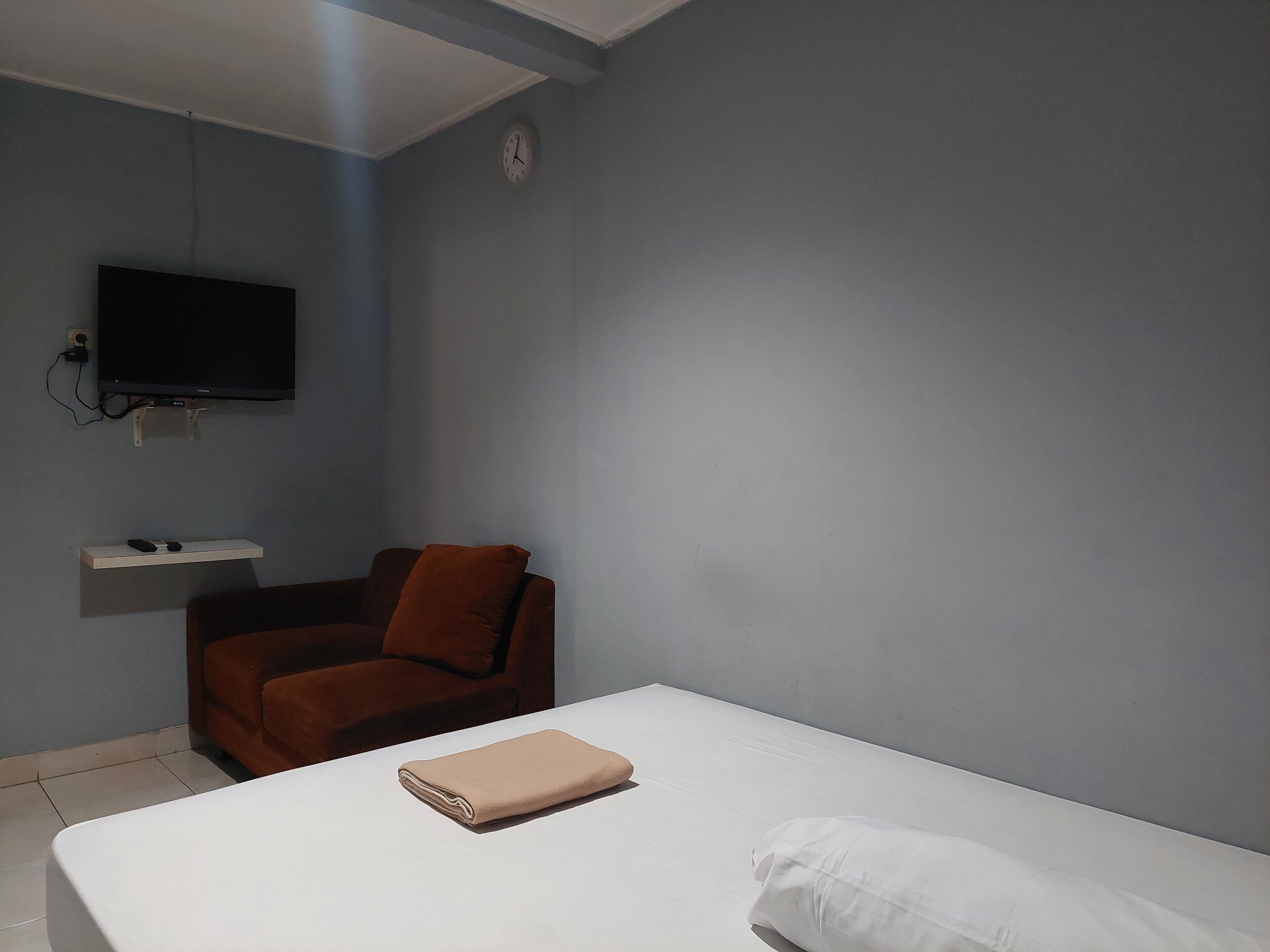 Una habitación con una cama, una silla marrón, un televisor montado en la pared y un estante pequeño.