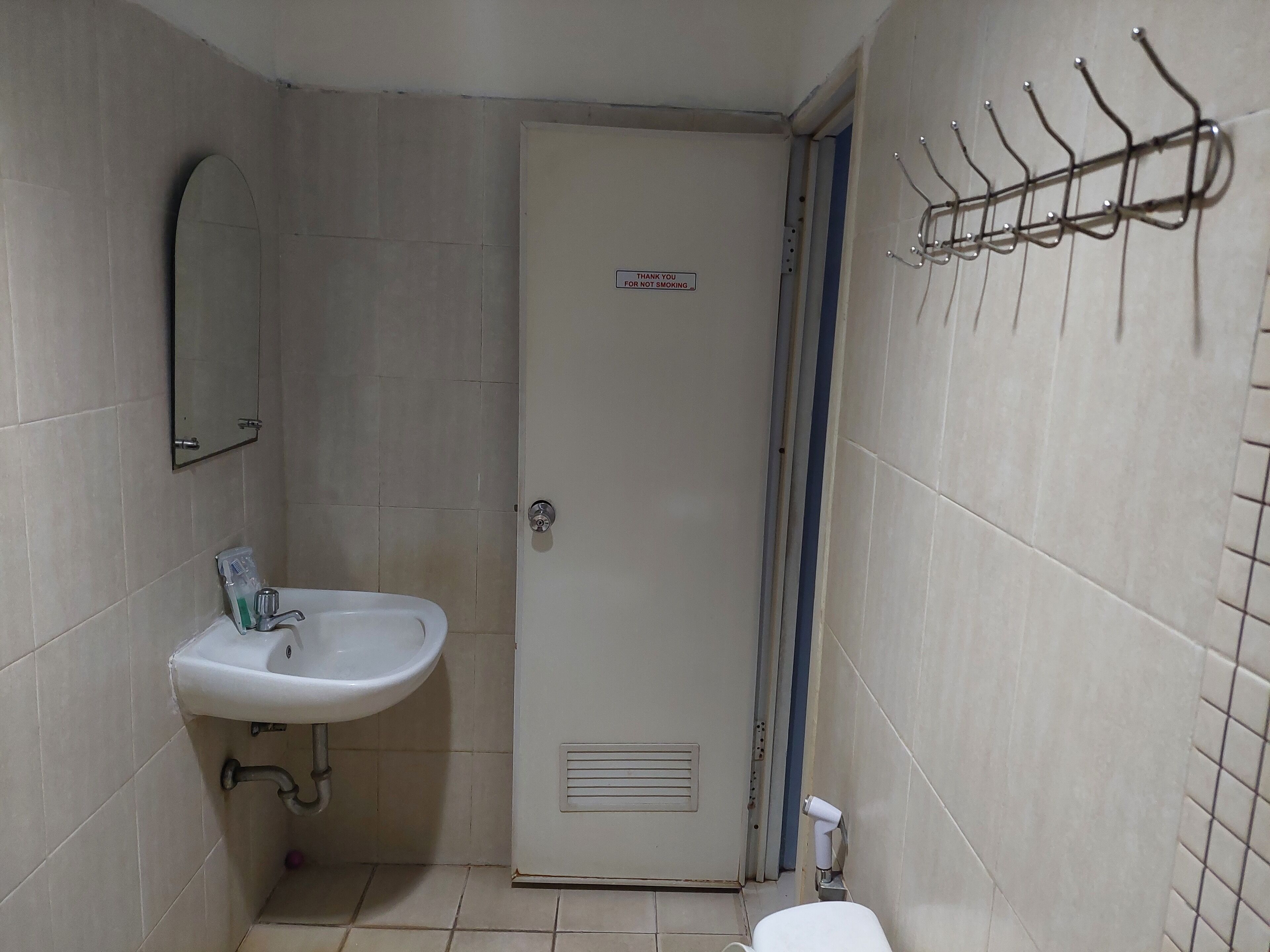 Une petite salle de bain avec un lavabo blanc, un miroir et une porte sur laquelle est apposée une pancarte « Interdiction de fumer ».