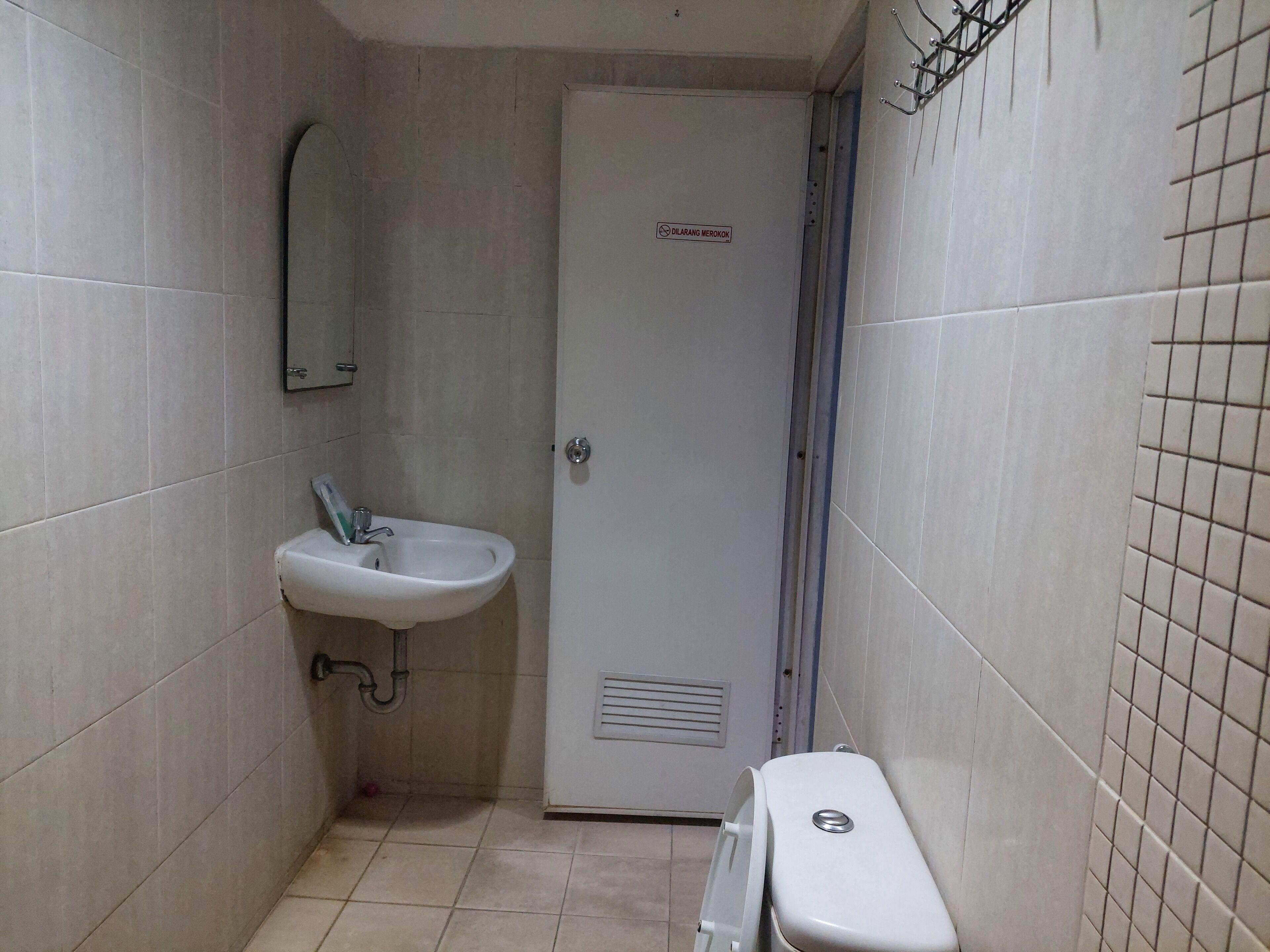 A small bathroom with a sink, toilet, and a door marked "DILARANG MEROKOK" (no smoking).